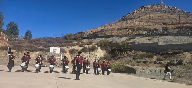 Banda de Guerra de uniforme guinda tocando en una explanada al pie de un cerro